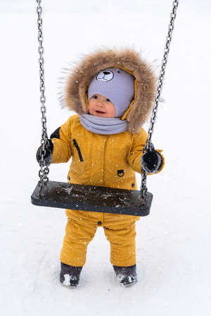 A happy toddler 12-17 months old in yellow winter clothes is holding onto a swing and trying to swing on them. A baby on a children's playground in winterの写真素材