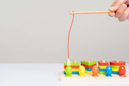A hand with a toy fishing rod with a magnet shows how to use a wooden sorter toy with colored puzzles and figures to simulate fishing. Children's educational gamesの写真素材