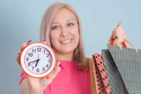 A pretty adult woman in her 40s with shopping bags stands on a blue background and holds a watch at arm's length. selective focusの写真素材