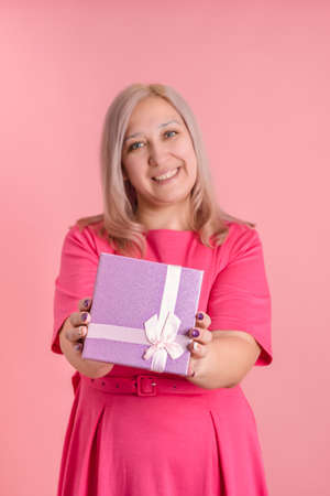 A blonde woman of 40 years old holds out a gift box standing on a pink background. selective focusの写真素材