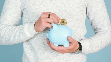 A man puts a gold coin bitcoin in a blue piggy bank