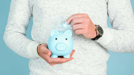 A man puts a silver coin in a blue piggy bank that he holds in his hands, close-upの写真素材