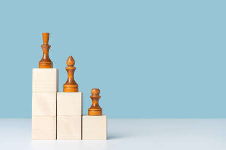 Wooden cubes arranged in the form of stairs and pyramids, on which black chess pieces are arranged in order of importance, a pawn officer and a queen. Layout for a business concept, copy space on a blue backgroundの写真素材