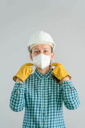 A young adult female construction worker in a protective medical mask stands on a gray background in a white hardhat adjusting a respirator on her face. vertical photoの写真素材