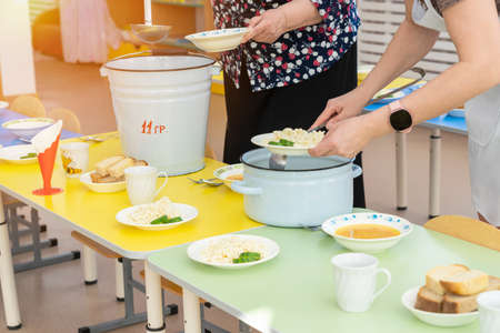 Nannies in kindergarten put food on plates and serve tables before lunchの写真素材