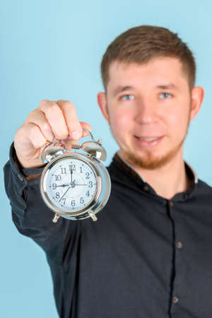 A young adult bearded man holds an alarm clock at arm's length standing on a blue background, selective focusの写真素材