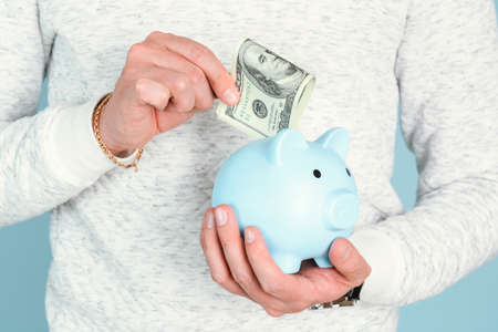 A man's hand puts a hundred-dollar bill in a blue piggy bank, close-upの写真素材