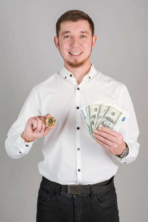 A businessman in a white shirt holds a bitcoin gold coin and a fan of paper dollar bills in his hand, vertical photoの写真素材