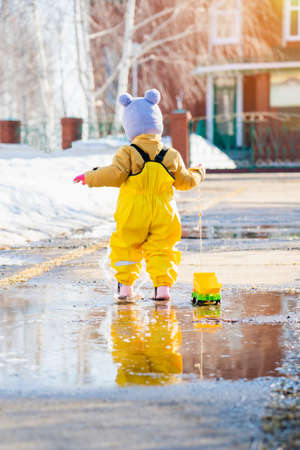toddler 2-3 years old dressed in yellow waterproof clothes runs through puddles with a toy truck tied on a rope. Spring 2022の写真素材