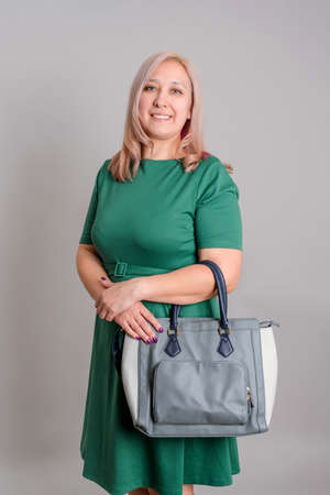 An adult blonde 40s woman in a green dress with a handbag stands on a gray background, vertical photoの写真素材