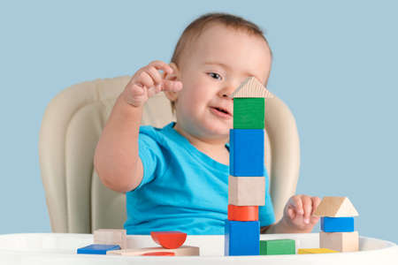 Smiling toddler 12-17 months old learns to build a tower of colored wooden cubes sitting on a child's chair, on a blue backgroundの写真素材