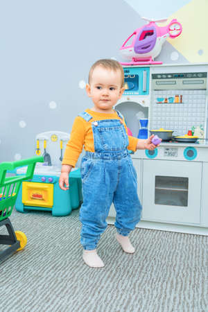 Cute child 2-3 years old in the playroom dressed in denim overalls playing with a plastic mug in the toy kitchenの写真素材