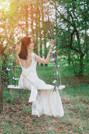 A beautiful girl in a dress with a cutout on the back is sitting on a rope swing in an apple orchard in springの写真素材