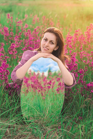 Portrait of a beautiful girl in a blooming meadow with an oval mirror in her hands in which purple wildflowers and the sky are reflectedの写真素材