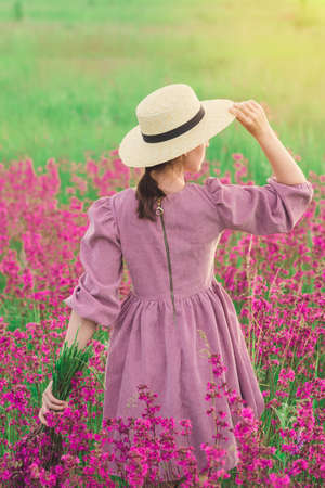 A girl in a straw hat and a purple dress walks through a field with blooming purple flowers at sunsetの写真素材