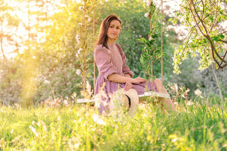 A beautiful confident girl in a purple dress poses on a rope swing in a blooming apple orchardの写真素材