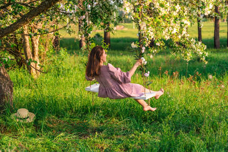 A brunette woman in a purple dress enjoys swinging on a rope swing in an apple orchard at sunsetの写真素材