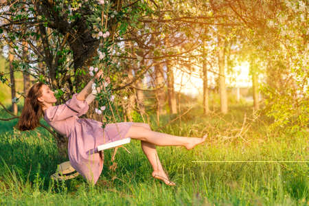 A brunette woman in a purple dress swings on a rope swing in an apple orchard at sunsetの写真素材