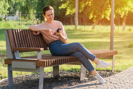 Beautiful brunette with a mobile phone sits on a bench in the parkの写真素材