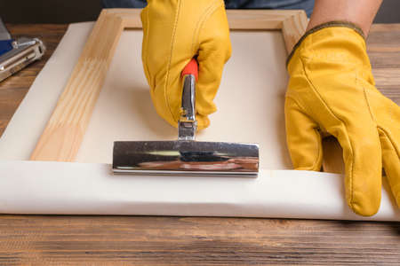 The artisan's hands in yellow protective gloves pull the canvas onto a wooden stretcher with tongsの写真素材