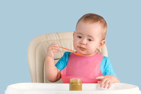 A baby 12-17 months old in a pink bib eats mashed broccoli with a spoon while sitting on a feeding chairの写真素材