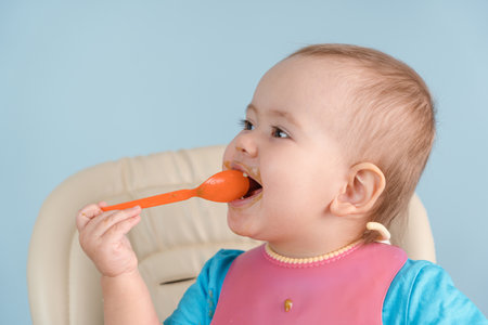 A happy smiling baby 12-17 months old in a pink bib puts a spoon with vegetable mashed broccoli in his mouthの写真素材