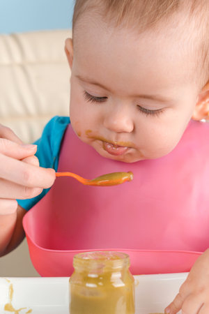 Mom feeds baby 12-17 months with a spoon of green vegetable puree, vertical photoの写真素材