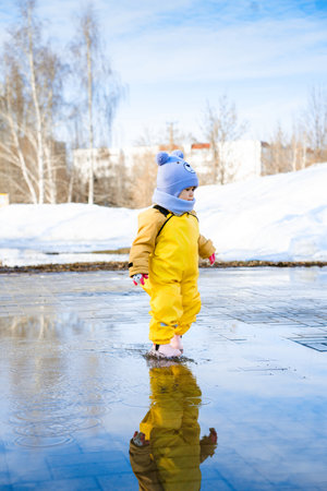 toddler 2-3 years old dressed in yellow waterproof clothes runs through puddles in a city park in the spring.の写真素材