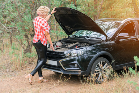 A blonde woman stands at the open hood of a car that broke down in the forestの写真素材