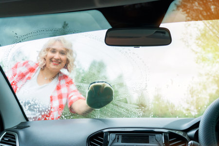 An adult blonde woman washes the windshield of a car with a washcloth, a view from inside the car interiorの写真素材