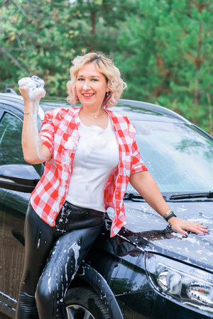An adult blonde woman sexually washes a car by spraying herself with soap foam, vertical photoの写真素材