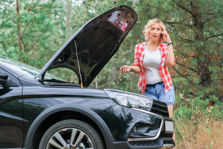 Breakdown on the road, a female driver uses a mobile phone while standing at the open hood of the carの写真素材