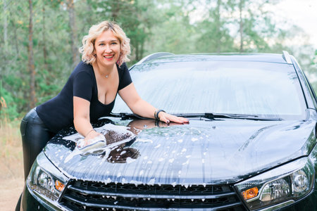 A middle-aged blonde woman washes a car standing at the hoodの写真素材