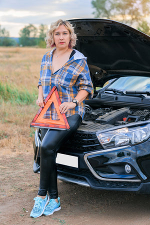 A woman holds a triangular emergency stop sign in her hand while standing at the open hood of a broken carの写真素材