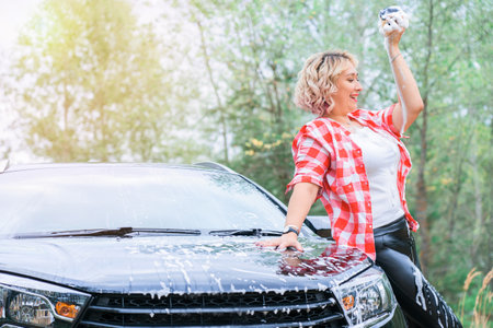 An adult blonde woman sexually washes a car by spraying herself with soap foamの写真素材