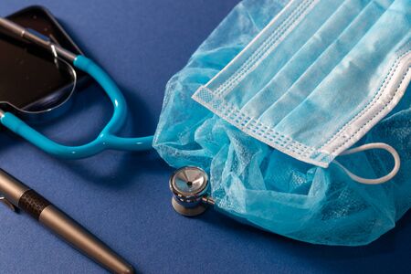 Neonatal blue stethoscope, medical cap and mask on blue dark backgrpund, pen and partial view of a cellular phone. Medical attention from specialist for premature and newborn babies concept.の写真素材