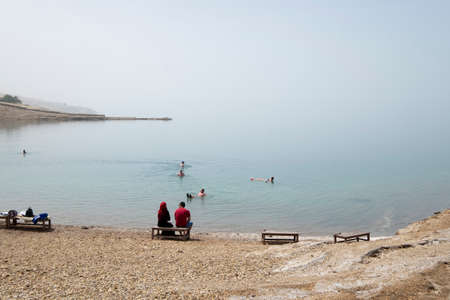 Tourists at the Dead Sea. Sitting on the wooden benches. People swimming in the sea. A landlocked salt lake between Israel and Jordan in southwestern Asia. Mountains in the background.のeditorial素材