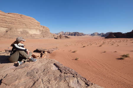 Wadi Rum, Jordan also known as Valley of the Moon. A female tourist sits on a rock enjoying the view of red dunes. A valley cut into the sandstone and granite rock in southern Jordan.のeditorial素材