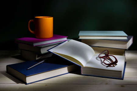 Stack of books with eye-glasses and a cup on a table. Low-key still-life photo, made on studio with black background and softbox.の写真素材