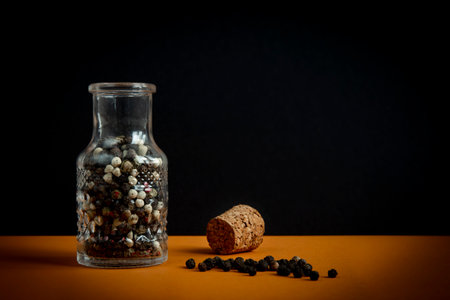 Black pepper in a glass jar. Spicy spice grains scattered on a table next to the open container.の写真素材