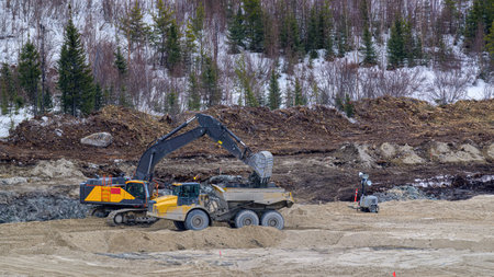 Excavator loading soil into a dumper at the foot of a hill in winterの写真素材