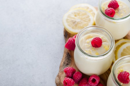 Three glass jars with lemon or vanilla dessert with raspberries and lemon slices on a wooden board, closeup, copy spaceの写真素材
