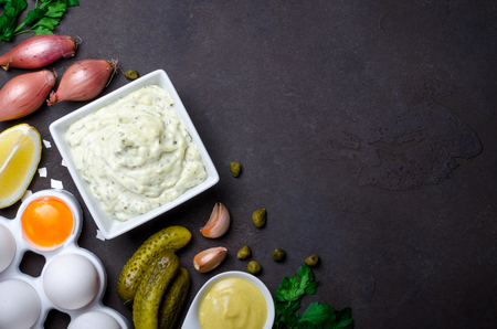 Homemade traditional french sauce remoulade in a white bowl with ingredients, lemon, eggs, shallot, pickles, parsley, mustard on a black dark stone slate background. Top view, horizontal, copy spaceの写真素材