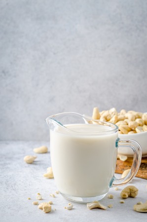 Cashew milk in a milk pitcher with cashew nuts on a light stone background. Front view, vertical image, copy spaceの写真素材