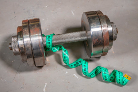 Steel collapsible dumbbell and green measuring tape around it on gray texture background. Workout and measurement concept. Sporty and healthy workout ideaの写真素材