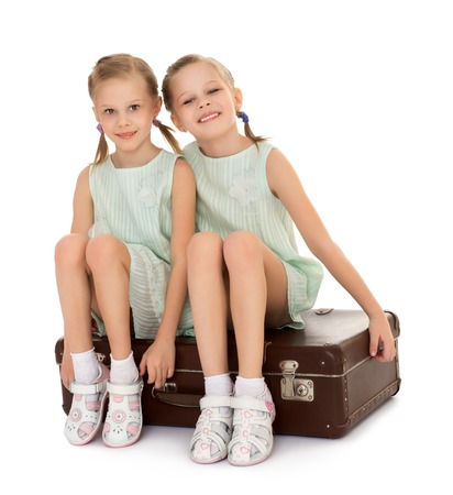 Beautiful , skinny small blonde girls sisters with thin pigtails on the head, in short summer dresses with short sleeves . Girl sitting on a Big old suitcase - Isolated on white backgroundの写真素材
