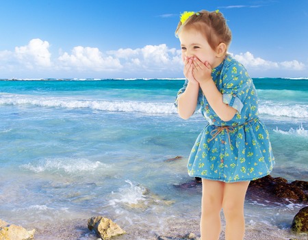 Cute little girl in a short blue dress terrified has closed hands the person. Close-up.On the background of white, sandy beaches, blue sky and clouds. The concept of a summer family vacation.の写真素材