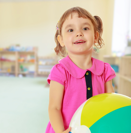 Cute little girl with pigtails on the head , in a pink dress. Girl holding a ball. Close-up.On blurred background the great hall of the kindergarten, with long racks where there are toys.の写真素材