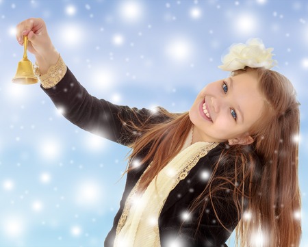 Joyful little long-haired girl with a white rose in her hair, rings the bell. Close-up.Blue Christmas background with white snowflakes.の写真素材