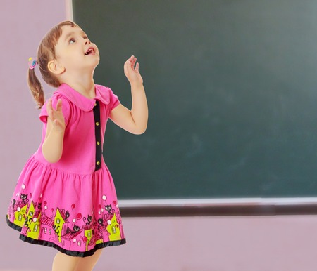 Pensive little girl with pigtails on the head , in a pink dress. The girl was looking at the top turned sideways to the camera.On a background of green, the school Board.の写真素材